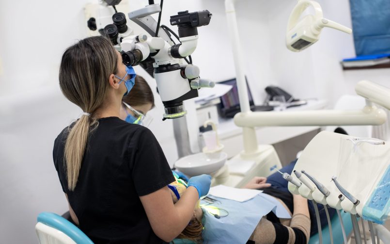 Tratament de canal asistat la microscop de către medicul stomatolog în partea stângă, pacient așezat pe scaun în partea de jos
